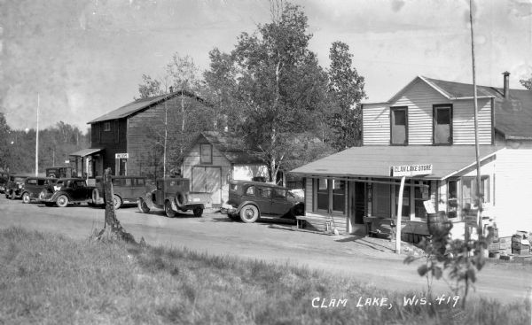 View across road of the Clam Lake Store on the right, a two-story building with a full front porch and a sign that reads “Clam Lake Store, General Merchandise." Along the road are parked automobiles. In the center is a garage and on the far left is another two-story building with a sign for "Rooms." (In a 1943 photograph this is labeled as the Chippewa Tavern.)
