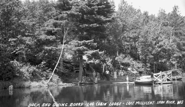 View from water of seven men, women and children standing on the shoreline at Log Cabin Lodge looking out over a diving board and dock on Lake Millicent. There are boats on shore and one boat tied up at the dock. In the background are trees and one man walking down a stone staircase to the shoreline.