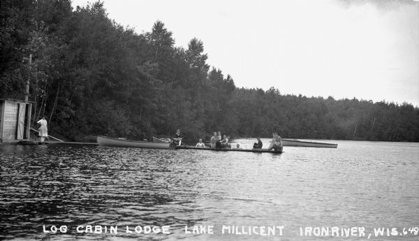 One woman stands on shore at Log Cabin Lodge looking at the photographer. Eight people sit on the dock, while two men are standing in Lake Millicent. Three boats are visible around the dock.