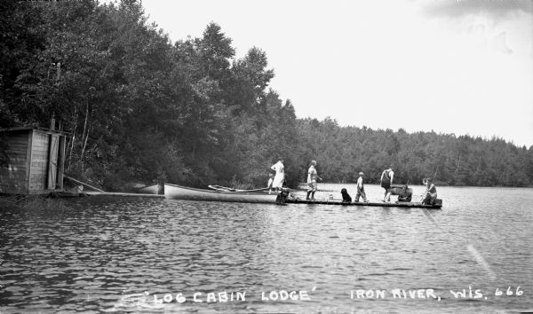 Nine people and one dog on the dock at Log Cabin Lodge on Lake Millicent. One man is swimming on the far side of the dock.  The shoreline and three boats are visible.