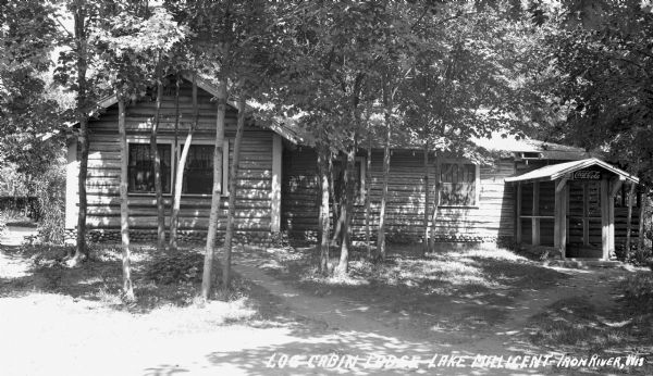 Exterior view of a one-story log cabin with stone foundation, the Log Cabin Lodge on Lake Millicent. There is a sign on the right side that says "Drink Coca-Cola."