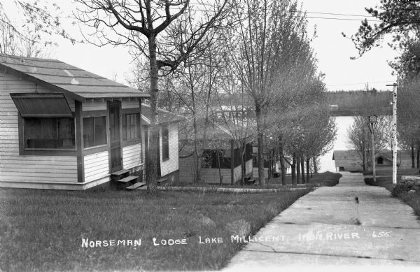 View of five cabins at Norseman Lodge on Lake Millicent. There are sidewalks between cabins, a birdhouse on the right, and the lake in the distance.