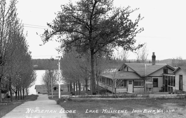 Exterior view of the Norseman Lodge with Lake Millicent visible in the background. The larger building on the right has a sign that says "Hotel." A bird house, sidewalks and the roof of another cabin are visible.