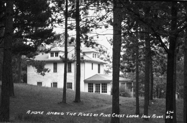 View through the pine trees of a two-story house that has a hipped roof with dormers and a one-story front porch at Pine Crest Lodge.