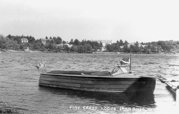 View of a wooden boat named the "Tip Top" tied to a dock on a lake at Pine Crest Lodge. In the background the opposite shoreline across the lake is visible with trees and buildings, including what appears to be two large barns.
