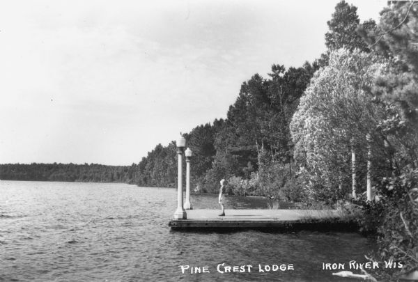 Child standing on a large dock with lights at Pine Crest Lodge. The lake and trees along the shoreline are also visible.