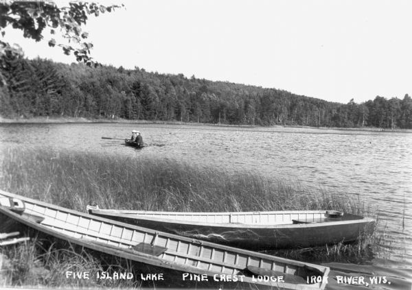View looking out over Five Island Lake at Pine Crest Lodge with two wooden row boats on shore in the foreground. Two people are rowing another boat in the lake.