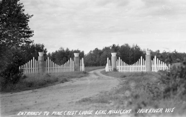 Entrance to Pine Crest Lodge. View of a dirt road and decorative fence with trees in the background. Caption reads: "Entrance to Pine Crest Lodge Lake, Millicent, Iron River Wis."