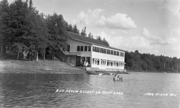 Three boys stand together in Deep Lake with four boats pulled up on shore behind them. On shore three women sit on a bench outside the Red Arrow Resort.