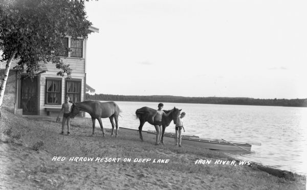 Three boys wearing swimsuits with two horses on the shore of Deep Lake with wooden row boats behind them. The Red Arrow Resort is visible on the left.