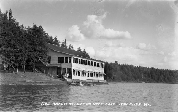 Three women, a dog, and two boys wearing swimsuits sit in front of the Red Arrow Resort building. Four wooden row boats are pulled up on the shoreline of Deep Lake and one boy, also wearing a swimsuit, sits on the end of one boat.