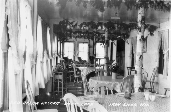 Interior view of the dining room at Red Arrow Resort on Deep Lake.  There are curtains on the windows and interior decorations.  Tables and chairs are set up, and the table in the foreground has place settings of dishes.
