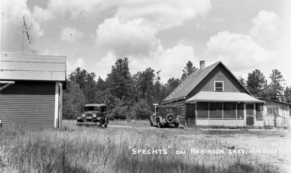 Specht's on Robinson Lake, view showing a house or cabin with gabled roof and front porch. On the left is another building with an American Flag. Two automobiles are parked in the center.