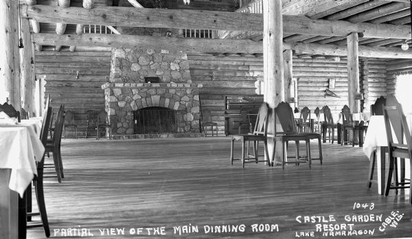 Interior view of part of the main dining room at the Castle Garden Resort on Lake Namakagon. The walls, columns and beams are logs. A large stone fireplace and a piano are on the back wall. Tables and chairs are set up around the room.