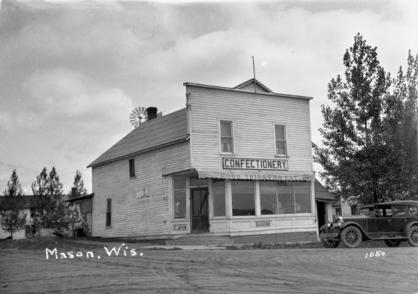 View of a two-story wooden building, the Mason Confectionery Store, with a sign that reads "Confectiony. Good Things to Eat." On the right is a parked automobile.