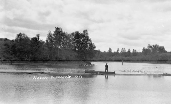 View from shoreline of a man standing in a boat fishing near Mason. The shoreline is visible in the background.