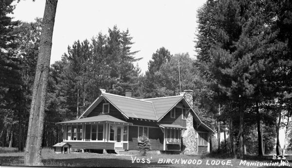 View of a two-story gabled roof lodge, Voss' Birchwood Lodge on Spider Lake. It has a stone chimney and a brick chimney, as well as a front porch and is surrounded by trees. There is a small doghouse on the left in front of the porch.
