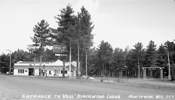 View of the entrance to Voss' Birchwood Lodge on Spider Lake. On the right are two birdhouses and a log entrance that says Voss' Resort. On the right is a service station with a sign that says "Rest Rooms, Tourist Information and Voss Birchwood."