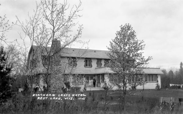 View across yard of three people standing on the porch of the two-story Northern Lakes Hotel on Rest Lake. There is a stone chimney and brickwork around the windows.