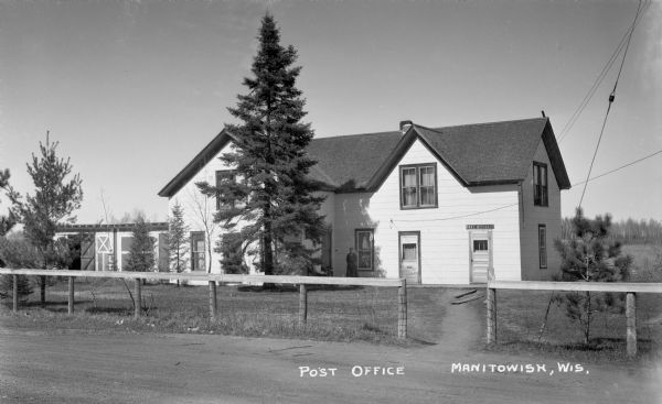 Woman standing outside of the two-story Post Office building in Manitowish. There is a road, fence and pine trees in the foreground.