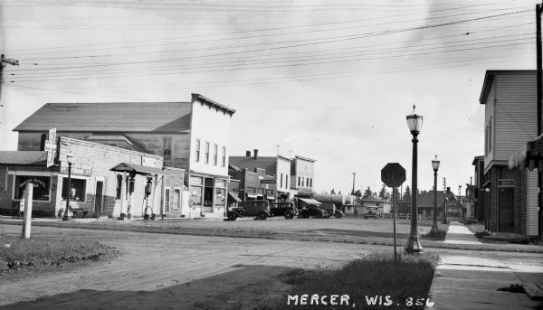 View looking at the automobiles parked in front of the stores along Main Street. Storefront signs on the left say: Mercer Auto Company, Mercer Garage, Goodyear, Drug Store, The Store, and Mercer Supply Co.  On the right is a Thru Traffic Stop sign and a sign for a Barber Shop.