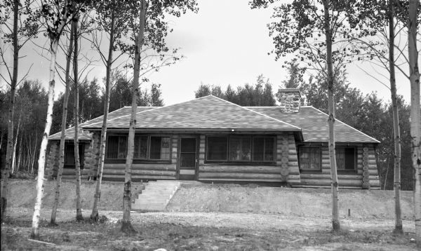 Exterior view of the front of a one-story log cabin home. There is a stone chimney on the right side.