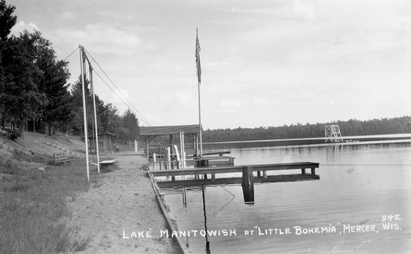 View down lake shoreline on Lake Manitowish at Little Bohemia. There are stairs coming down to the lake, and a bench, as well as docks, a flag pole and a diving raft are out in the lake.