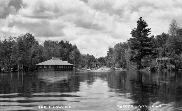 View across water of The Flowage showing the Mercer Fish Hatchery building on the left. Two people are in a boat on the far left, a bridge is in the center background and another building is on the right.