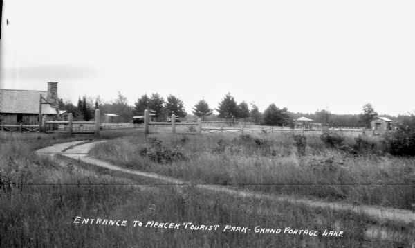 View of the road and entrance to Mercer Tourist Park on Grand Portage Lake showing the fences, road and buildings. The larger building on the left has a stone chimney.