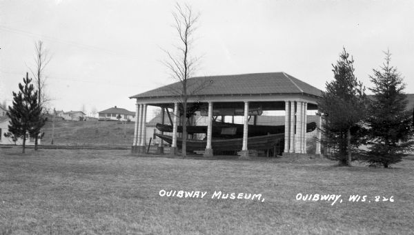 Canoes and wooden boats under a shelter at the Ojibway Museum. Other buildings, probably cottages, are visible in the background.