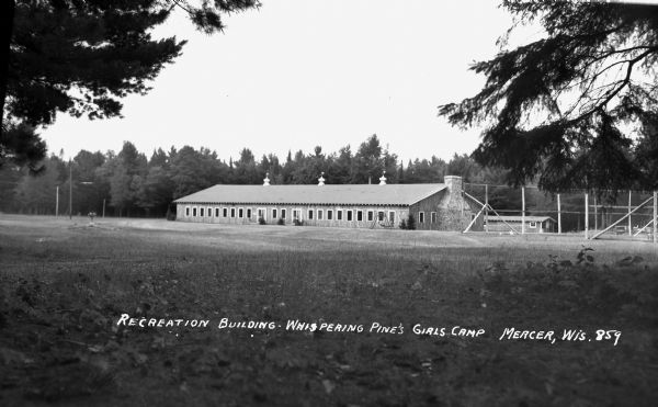 View across field of a large one-story wooden building with a large stone chimney on the right side. This is the Recreation building at Whispering Pines Girls Camp.