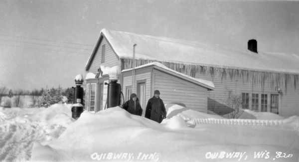 Two men standing outside of a one-story building, the Ojibway Inn, with large snow drifts around the building. One chimney is visible, and icicles hang down from the roof.