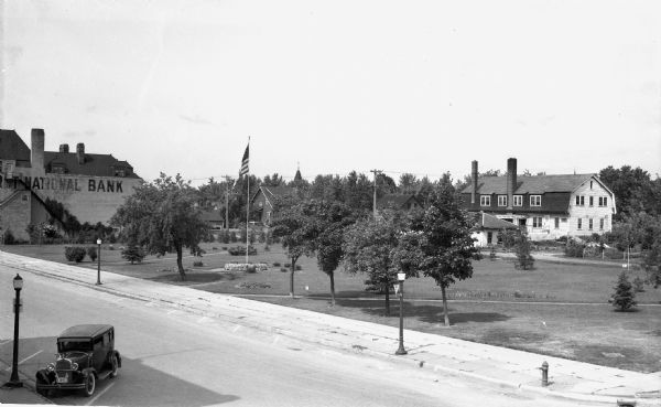 Elevated view of one automobile parked on Main Street near a park (the Legion Memorial Park), trees, an American flag, street lights, and various buildings, including one that says "First National Bank."
