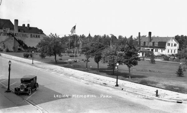 Elevated view of one automobile parked on Main Street. The Legion Memorial Park is across the street. There is a hospital building in the background. Trees, an American flag, street lights and various buildings surround the park, including a building that says "First National Bank."