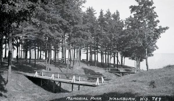 View of two wooden foot bridges in Memorial Park. In the background are the waters of Chequamegon Bay in Lake Superior. Picnic tables are among the trees. Memorial Park opened to the public in 1920.