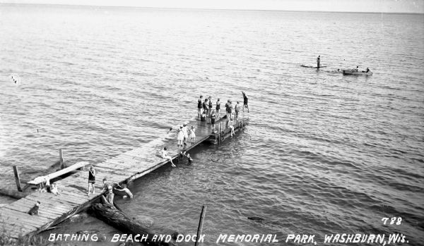 Boys and girls wearing bathing suits standing, swimming, or diving off of a large wooden dock into Chequamegon Bay, Lake Superior on the beach at Memorial Park. Two dogs sit on the dock. In the distance there is a man standing on a floating raft, another person swimming, and two men rowing a wooden boat.