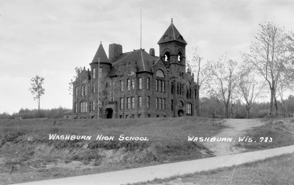 Exterior view of the Washburn High School, a two-story cut stone building with an attic and raised basement in the Romanesque architectural style. There is a square three-story tower on the right facade with the bell tower at the top. A round tower is also visible at the center of the left facade. A path, trees and grass surround the school building.