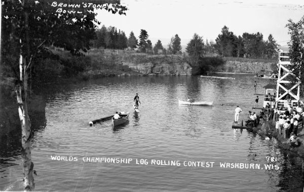 View of the World's Championship Log-Rolling Contest, Brownstone Bowl. There are two rowboats in the water. Two men stand on a rolling log in the middle of the water while a crowd of spectators stands on shore. There is a dock and diving boards right on the shoreline.