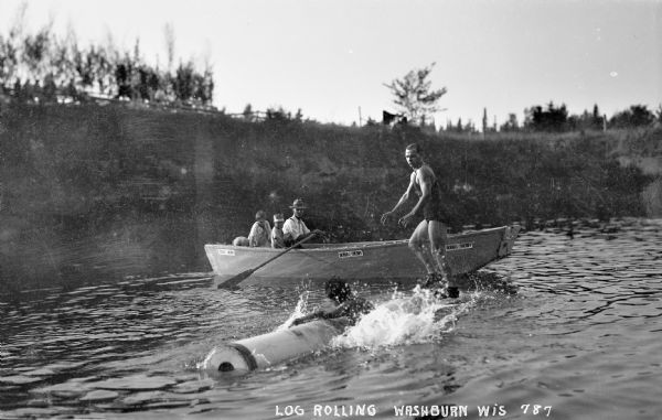 View from water of log rolling with one man in the water holding onto a log, and another man standing on a rolling log. Behind the log is a wooden rowboat that says "Demars for Sheriff," with two children and a man rowing the boat.