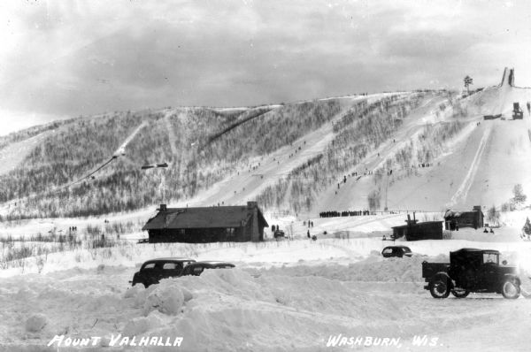 View of Mount Valhalla during the winter near Washburn. There is a building and a few automobiles parked at the bottom of a large hill covered in snow. The hill has various tracks for downhill skiing.