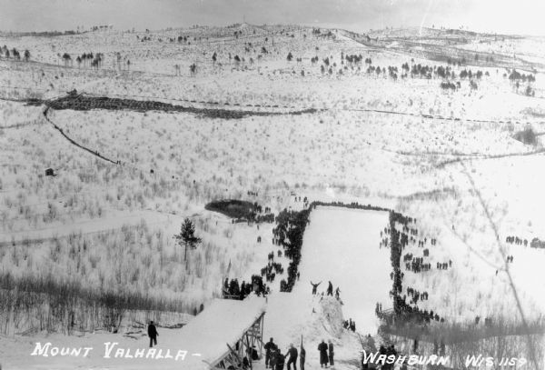 View from the top of a ski hill jump on Mount Valhalla near Washburn. Crowds of people line the edges of the ski jump area at the bottom of the hill.