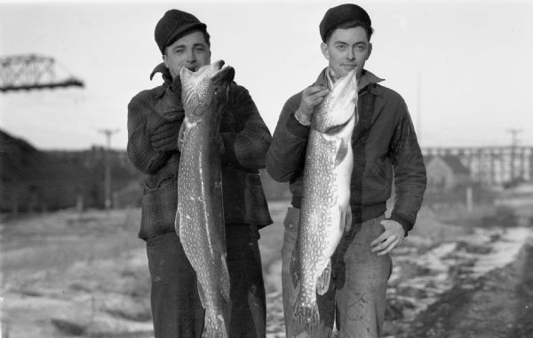 Two men wearing winter hats and coats holding up two large northern pike.