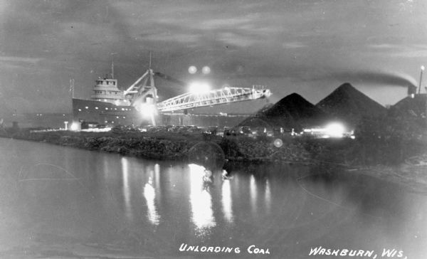 Unloading coal from a large ship in Lake Superior onto land near Washburn. There is a large conveyor that has lights glowing in the dark sky. A large pile of coal is on the right.