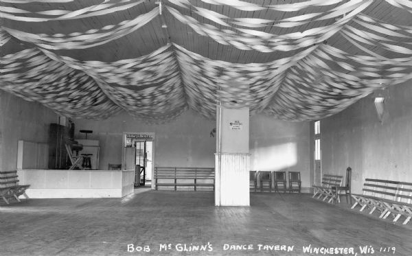 Interior view of Bob McGlinn's Dance Tavern showing the wooden dance floor with chairs and benches along the walls. In the back left corner is a stage. There are decorative streamers hanging from the ceiling and a pillar in the center of the room has a sign that reads "No Smoking While Dancing Please."