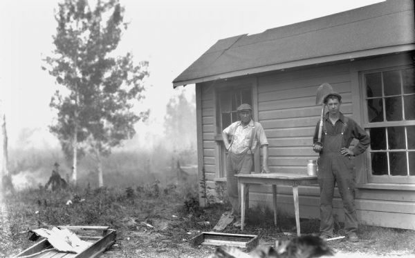 Two men wearing hats are standing outside of a cabin. One man, wearing suspenders, is leaning on a table. The other man, wearing overalls, is holding a shovel.