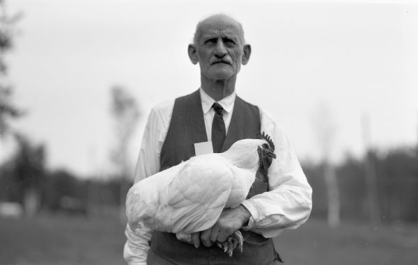 Man wearing a vest and tie stands holding a rooster at the Iron County Fair.