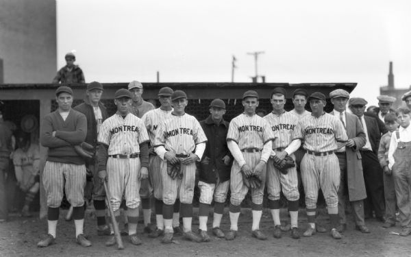 Group portrait of the Montreal men's baseball team outside of the baseball dug out. All of the men are wearing hats, a few have baseball gloves, and one man is holding a baseball bat.