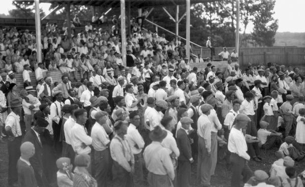 Crowd of men, women, and children standing in front of, or sitting in, the grandstand watching a baseball game at the Iron County Fair.