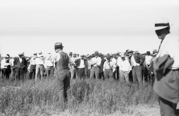 View of one man standing in the middle, while other men stand in a crowd, in an oat field. Most of the men are wearing suits and hats.