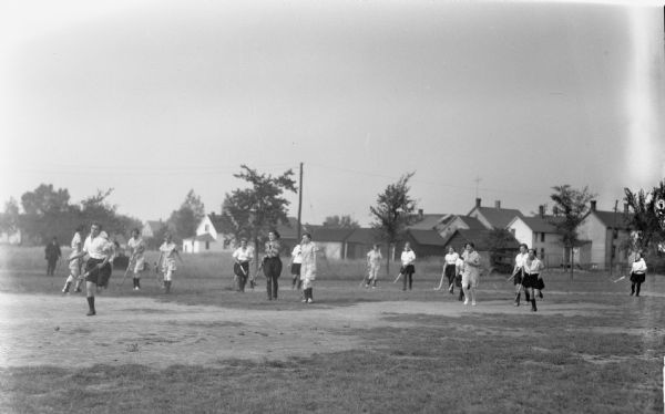 Girls running around on a field holding hockey sticks playing field hockey, possibly in Iron County. In the background are houses and trees.